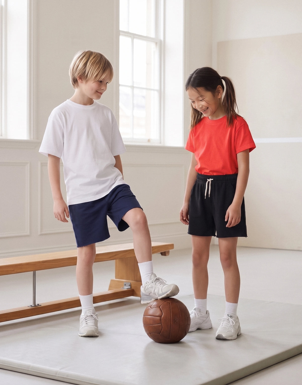 Two children standing on a gym floor mat indoors, one in a white t-shirt and navy shorts kicking a brown ball, and the other in a red t-shirt and black shorts looking on. No visible brand logos.