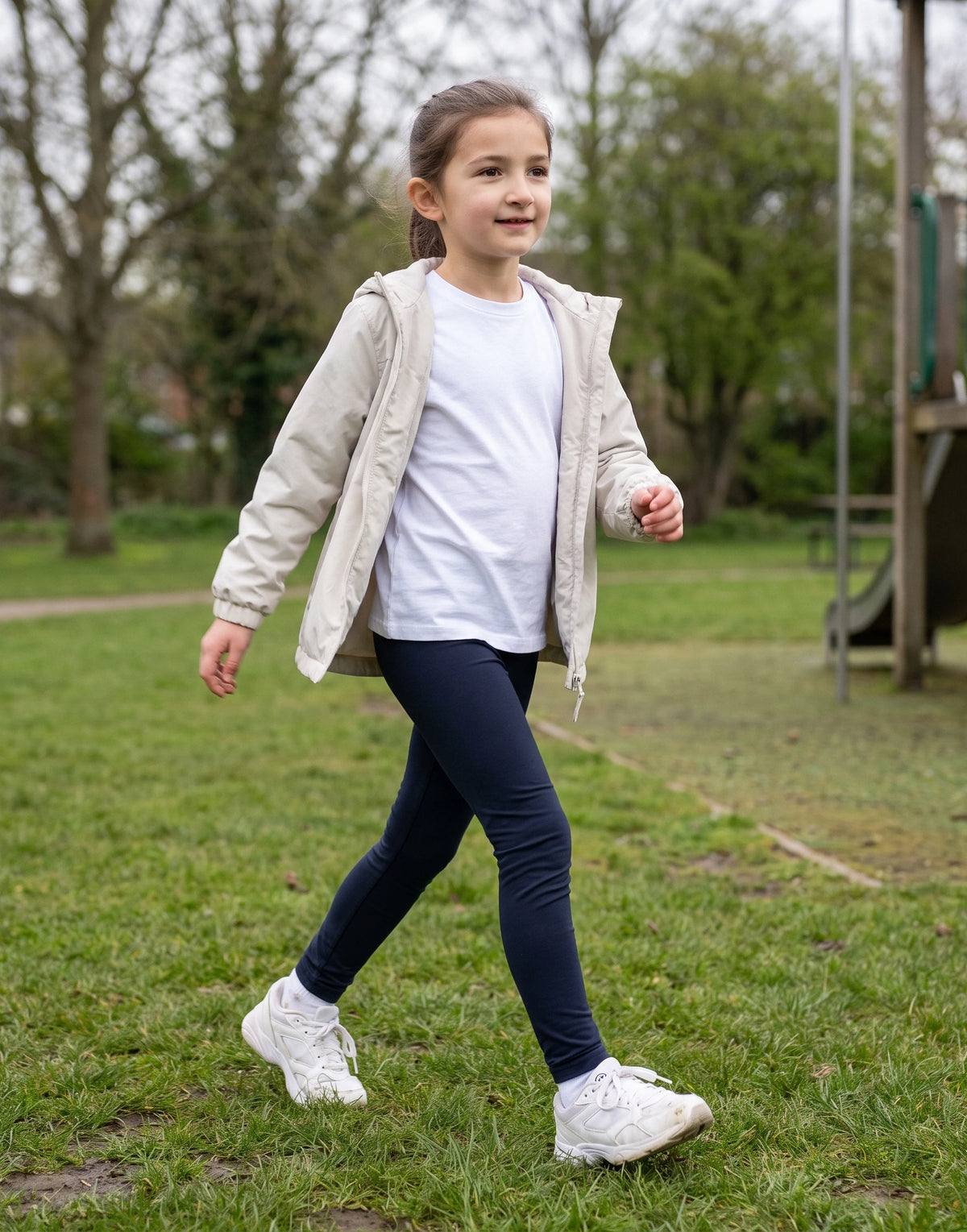 A young girl walking on a grassy park path wearing a light beige hooded jacket over a white t-shirt, dark navy leggings, and white sneakers.