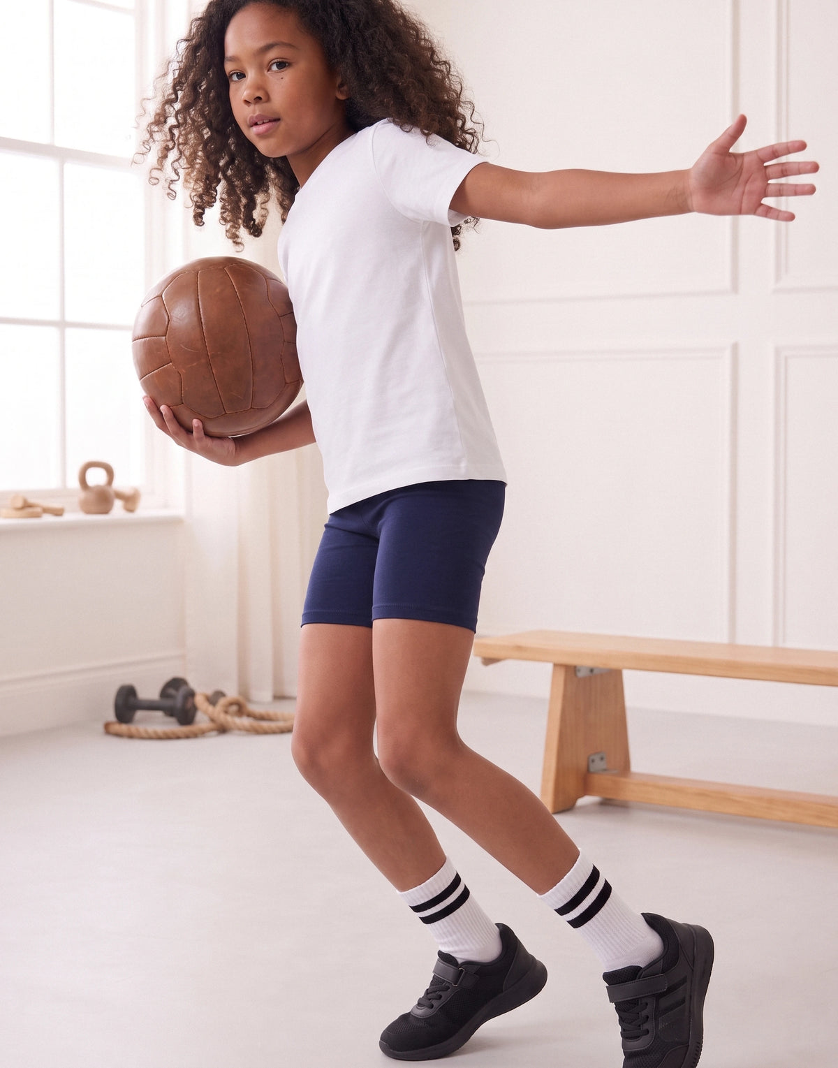 A young girl with curly hair is captured mid-action in a bright indoor room, wearing a white t-shirt, navy blue athletic shorts, white socks with black stripes, and black sneakers while holding a brown leather basketball.