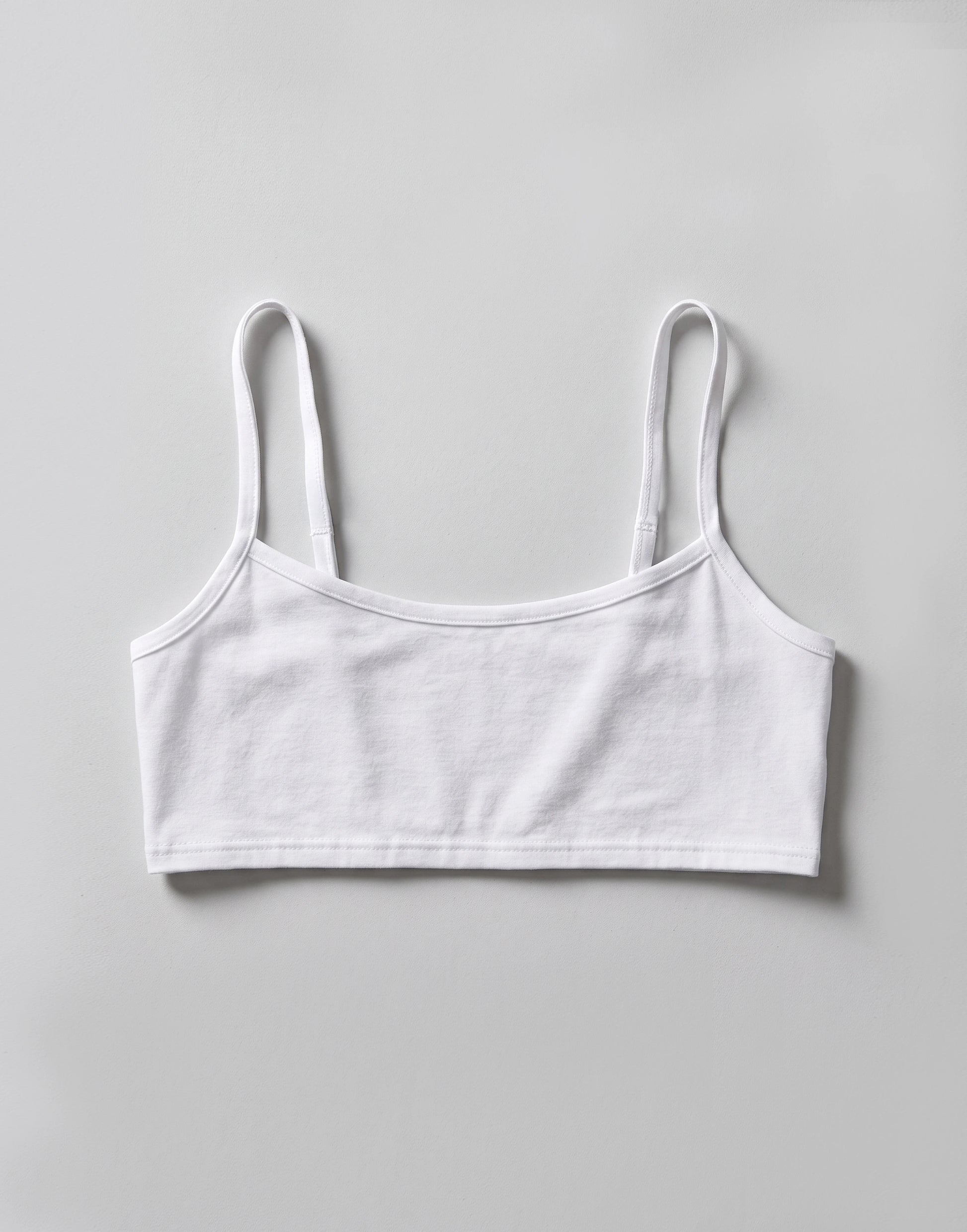A white cotton crop top with thin shoulder straps, laid flat against a light gray background.