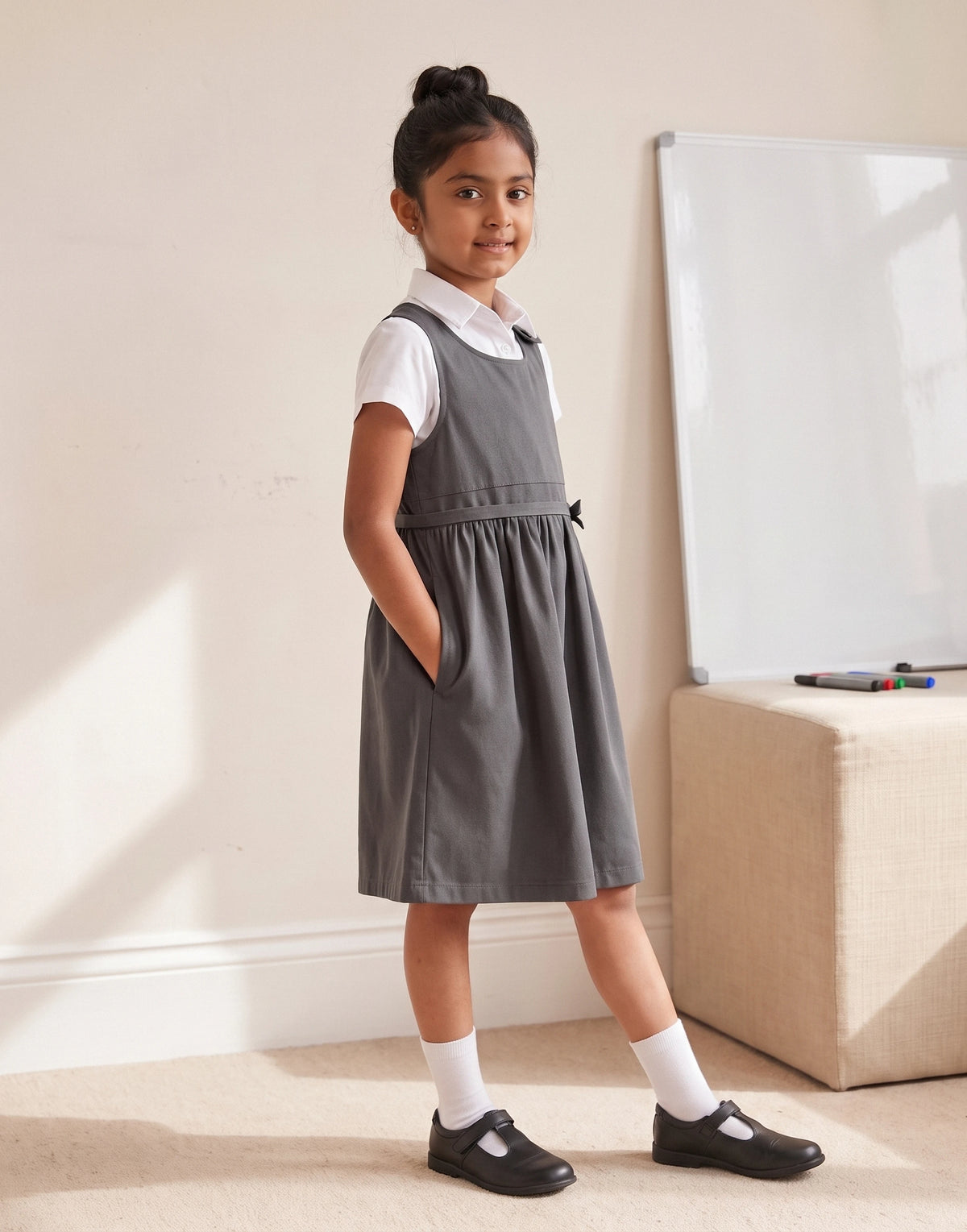 A young girl stands in a room wearing a gray pinafore-style school dress over a white short-sleeve collared shirt, with white socks and black Mary Jane shoes. A beige ottoman and a whiteboard are visible in the background.