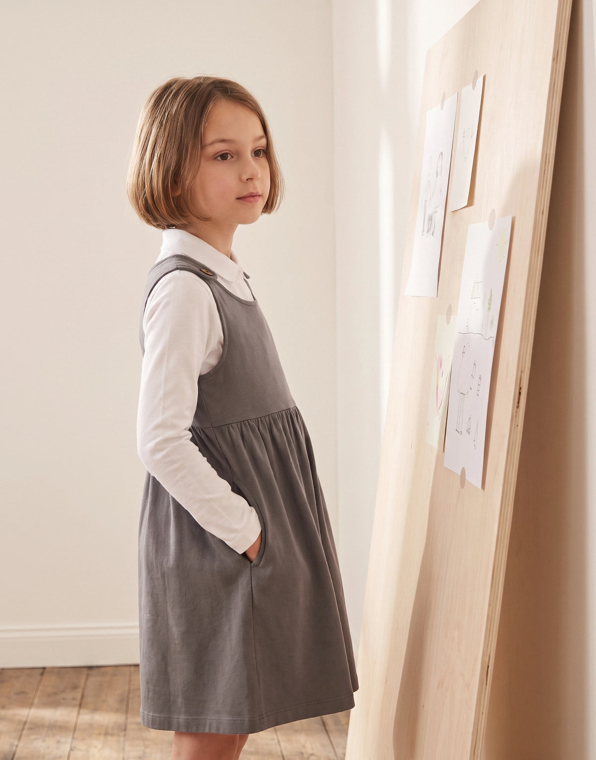 A young girl with short brown hair stands in profile wearing a gray pinafore-style dress over a white long-sleeve collared shirt. She is posed indoors next to a light wood board with pinned papers.