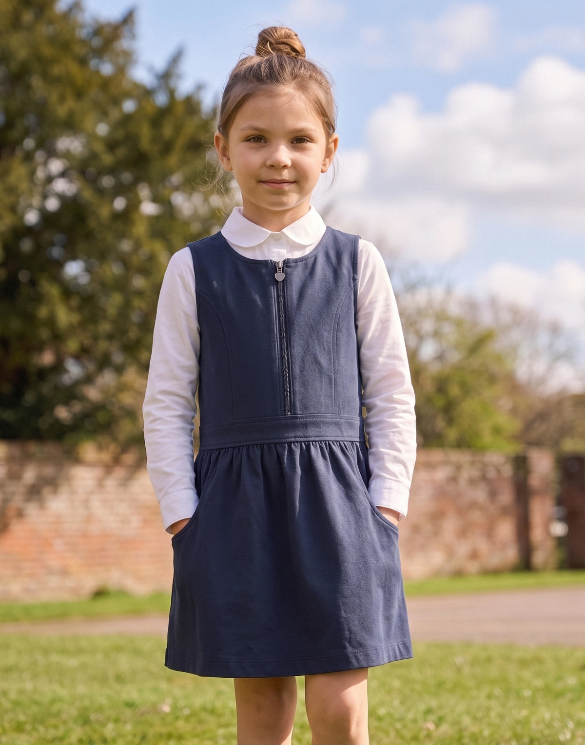 A young girl with her hair in a bun stands outdoors wearing a navy blue pinafore-style dress over a white long-sleeve collared shirt. The photo is taken in a grassy park area with trees and a brick wall in the background.