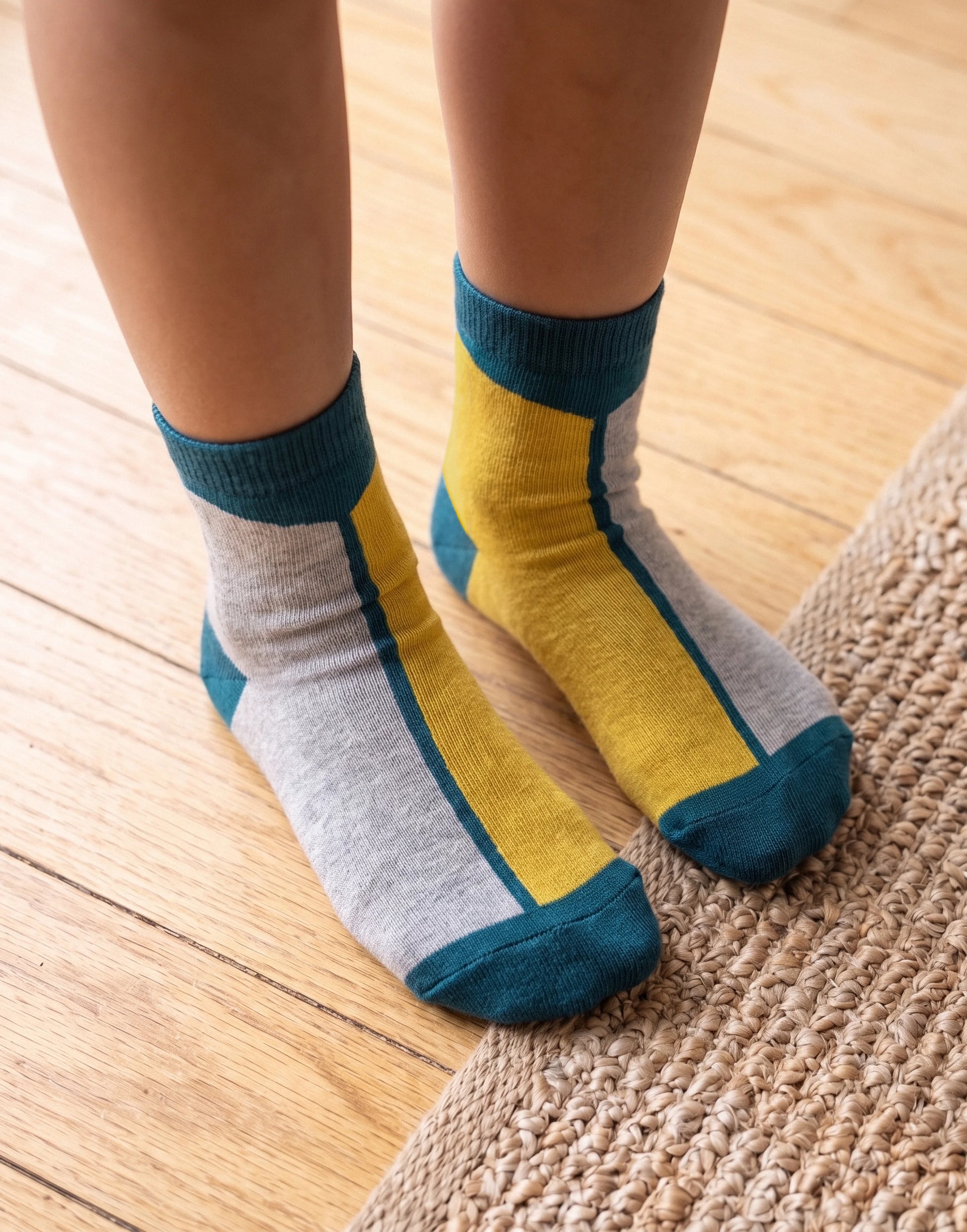 A close-up of a child's feet wearing striped ankle socks with gray, mustard yellow, and teal panels, standing on a light wood floor next to a woven rug.