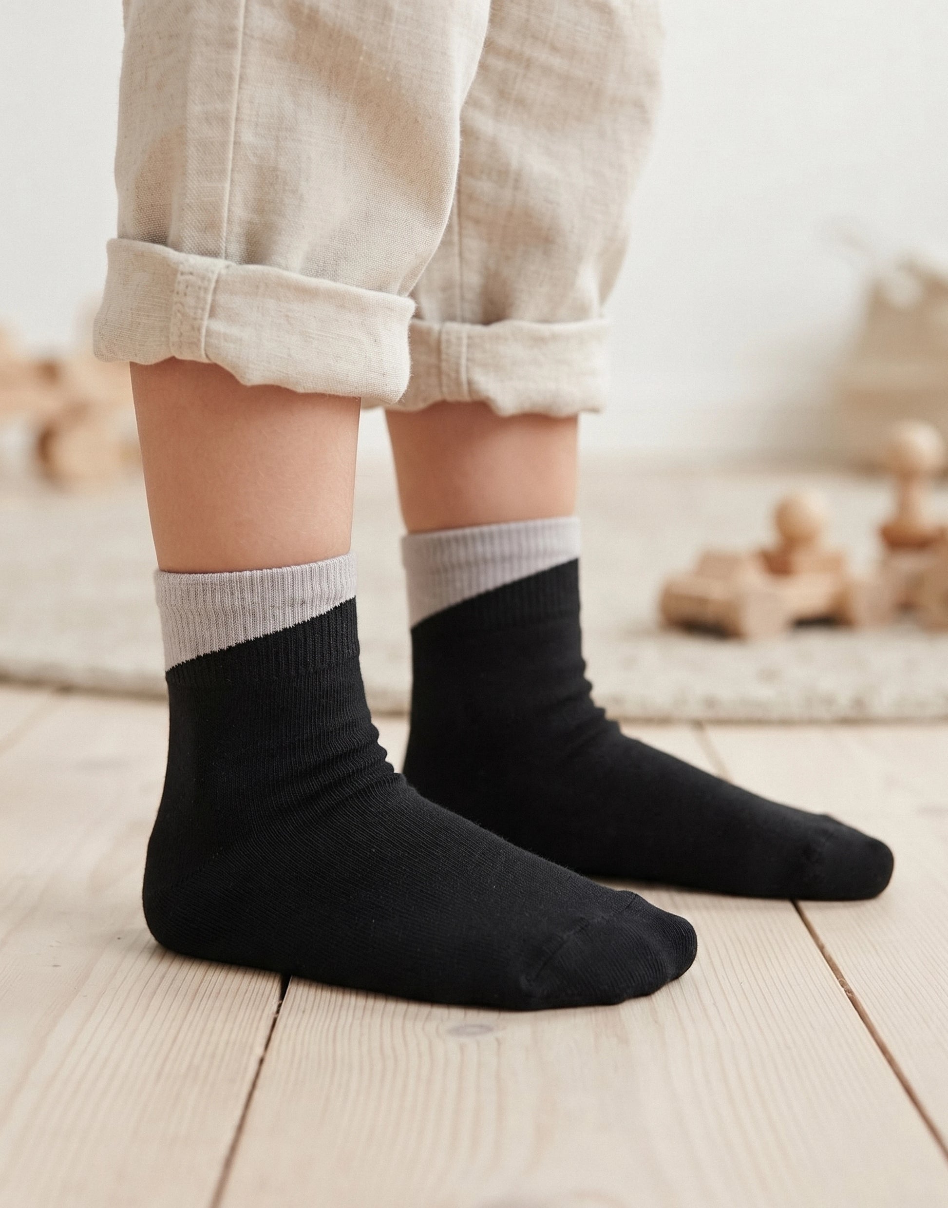 A close-up of a child's feet wearing black ankle socks with a light gray ribbed cuff, paired with rolled-up beige linen pants on a light wooden floor.
