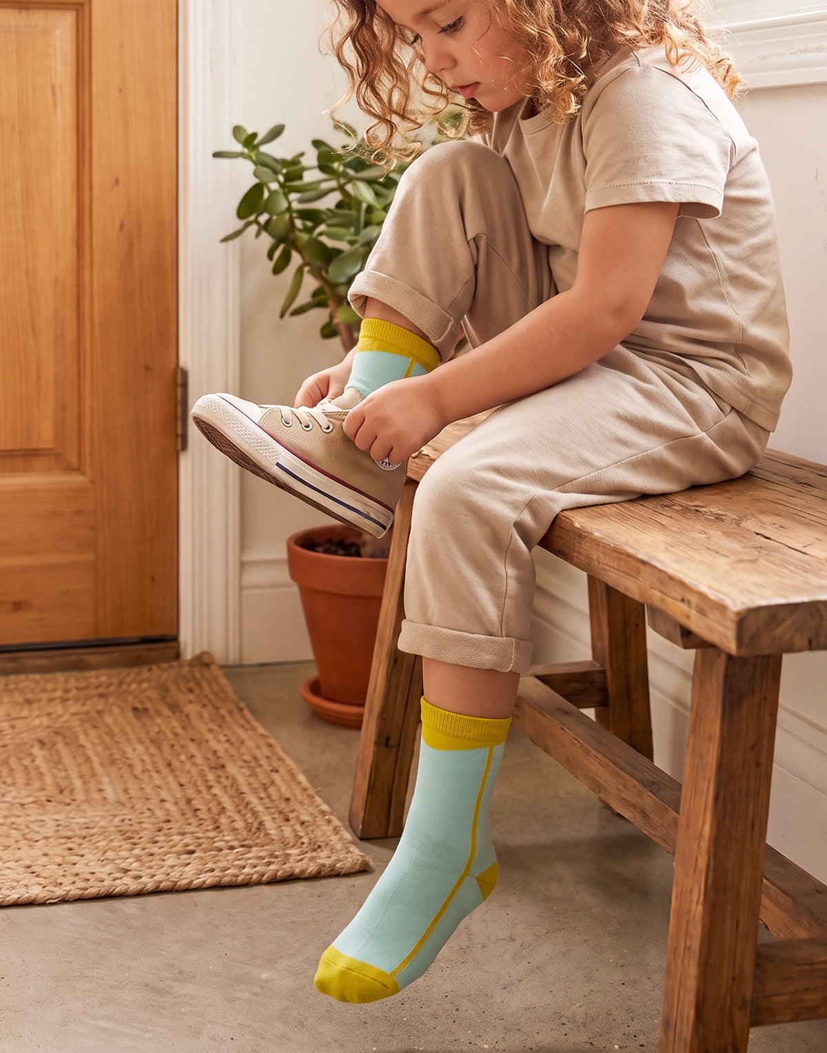 A child sitting on a wooden stool indoors, putting on a light blue sock with yellow trim and a yellow toe cap. The child is wearing a beige short-sleeve top and matching beige pants, and the scene includes a potted plant, door, mat, and concrete floor.