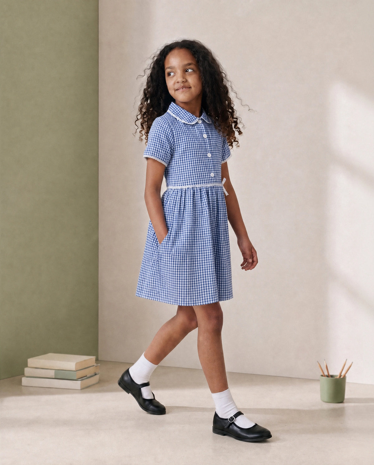A young girl with curly dark hair models a blue and white gingham short-sleeve dress with a white collar and button details, styled with white socks and black Mary Jane shoes in a minimalist studio setting.