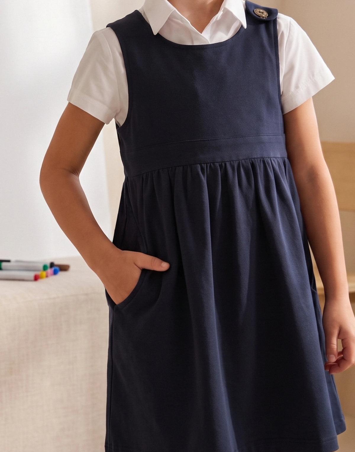A child wearing a navy blue pinafore dress over a white short-sleeve collared shirt, standing indoors with one hand in the dress pocket.