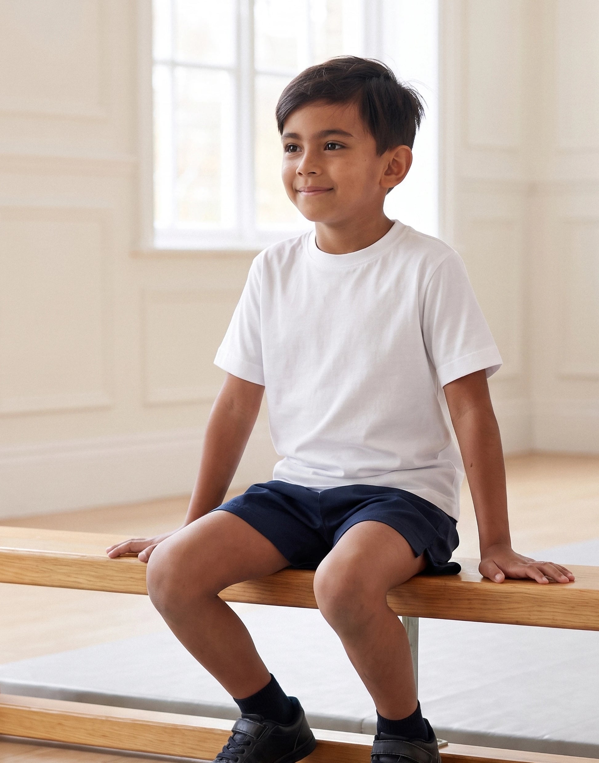 A young boy sitting on a wooden ballet barre in a bright, neutral room, wearing a plain white short-sleeve crewneck t-shirt and navy blue shorts.