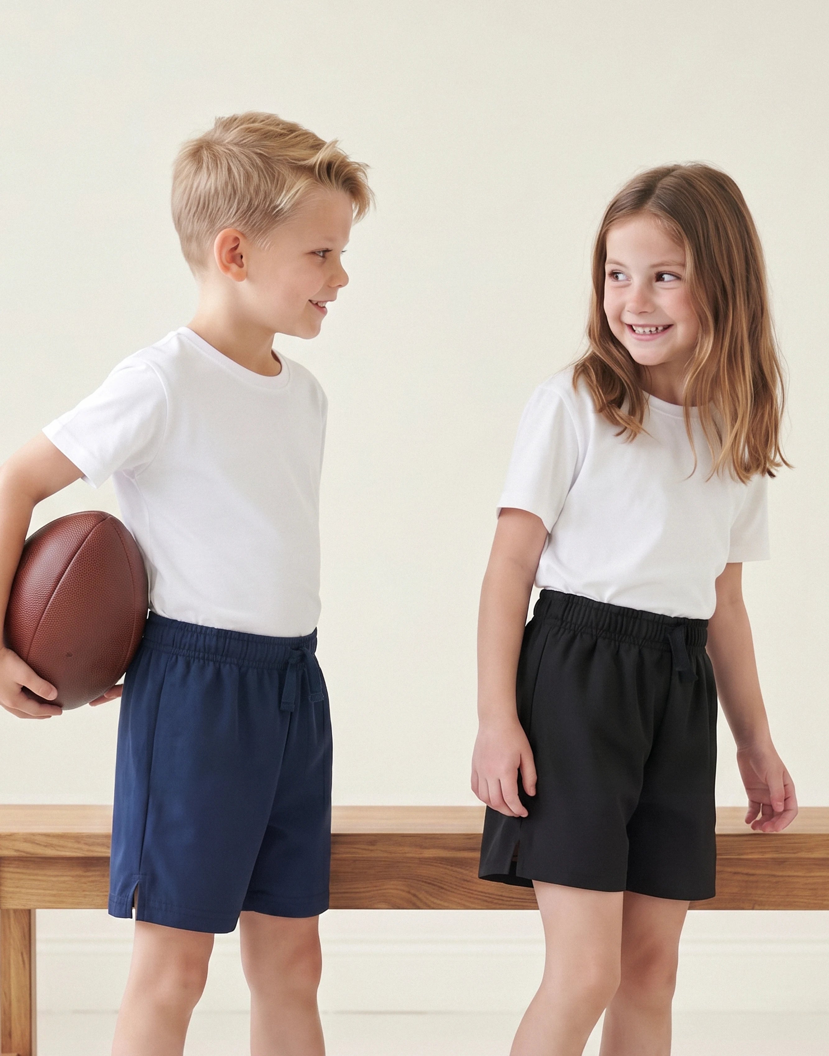 Two children standing indoors wearing plain white short-sleeve T-shirts and contrasting shorts, one in navy blue and one in black. The child on the left holds a brown football.